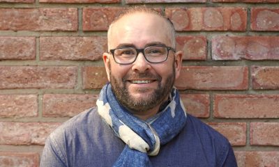 A white man with a beard and glasses smiles in front of a brick wall.
