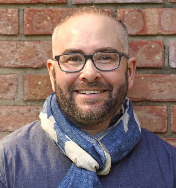 A white man with a beard and glasses smiles in front of a brick wall.